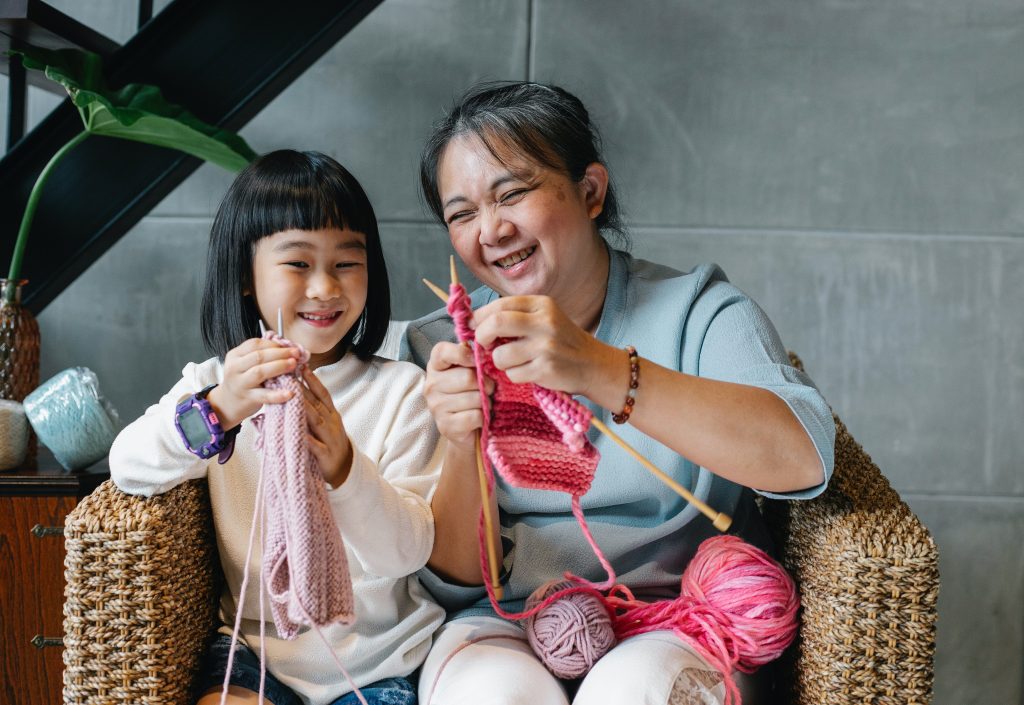 A young girl and woman share a chair; both are knitting and smiling