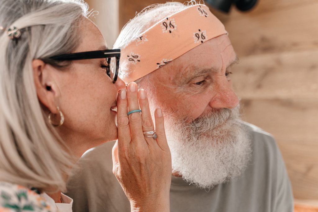 An older woman whispers in an older mans ear, she is holding her hand up to her mouth to keep her voice from being heard
